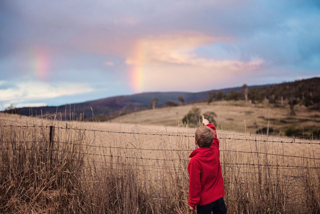 A boy of around 10 years old points at a rainbow and interesting cloud formation in the sky. He is standing in a hilly field with dry grass and a barbed wire fence.